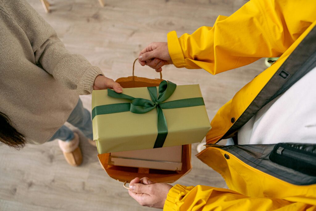 A courier in a yellow jacket hands over a gift in a paper bag indoors.